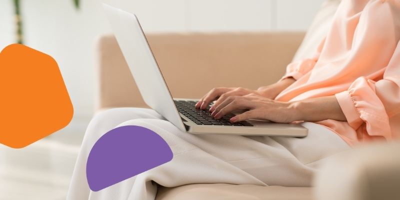 A woman types on a laptop while sitting on a sofa. She is wearing a pink blouse and beige pants. Her face can't be seen.