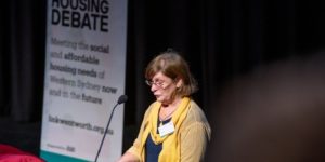 Mary, a middle-aged woman with mid-length brown hair delivering her speech on stage at the Big Housing Debate. She is wearing glasses and a yellow blouse.
