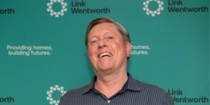 Mark, a middle-aged man smiles in front of a teal media wall with small Link Wentworth logos and the words, Providing Homes, Building Futures. He is wearing a dark dress shirt and his hair is combed back.